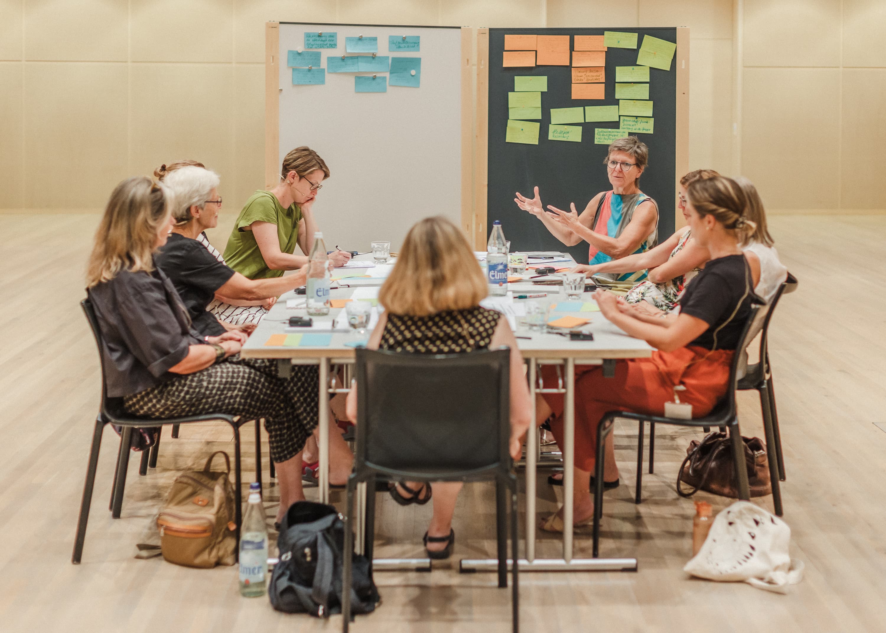 Eine gruppe von 9 Personen sitzt um einen Tisch und diskutiert angeregt. Im Hintergrund sieht man viele Post-its an einer Wand.