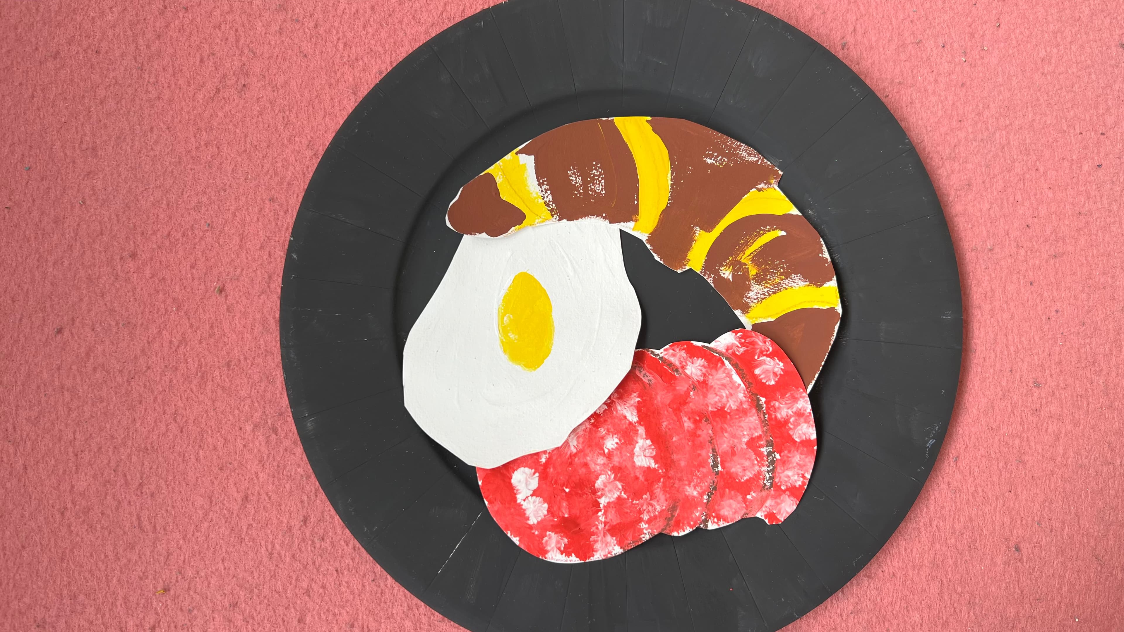 Auf einem lachsfarbenen Hintergrund ist ein schwarzer Teller platziert. Auf dem Teller befindet sich ein Spiegelei, ein Croissant und ein paar Scheiben Wurst. Alles ist aus Papier und bemalt.