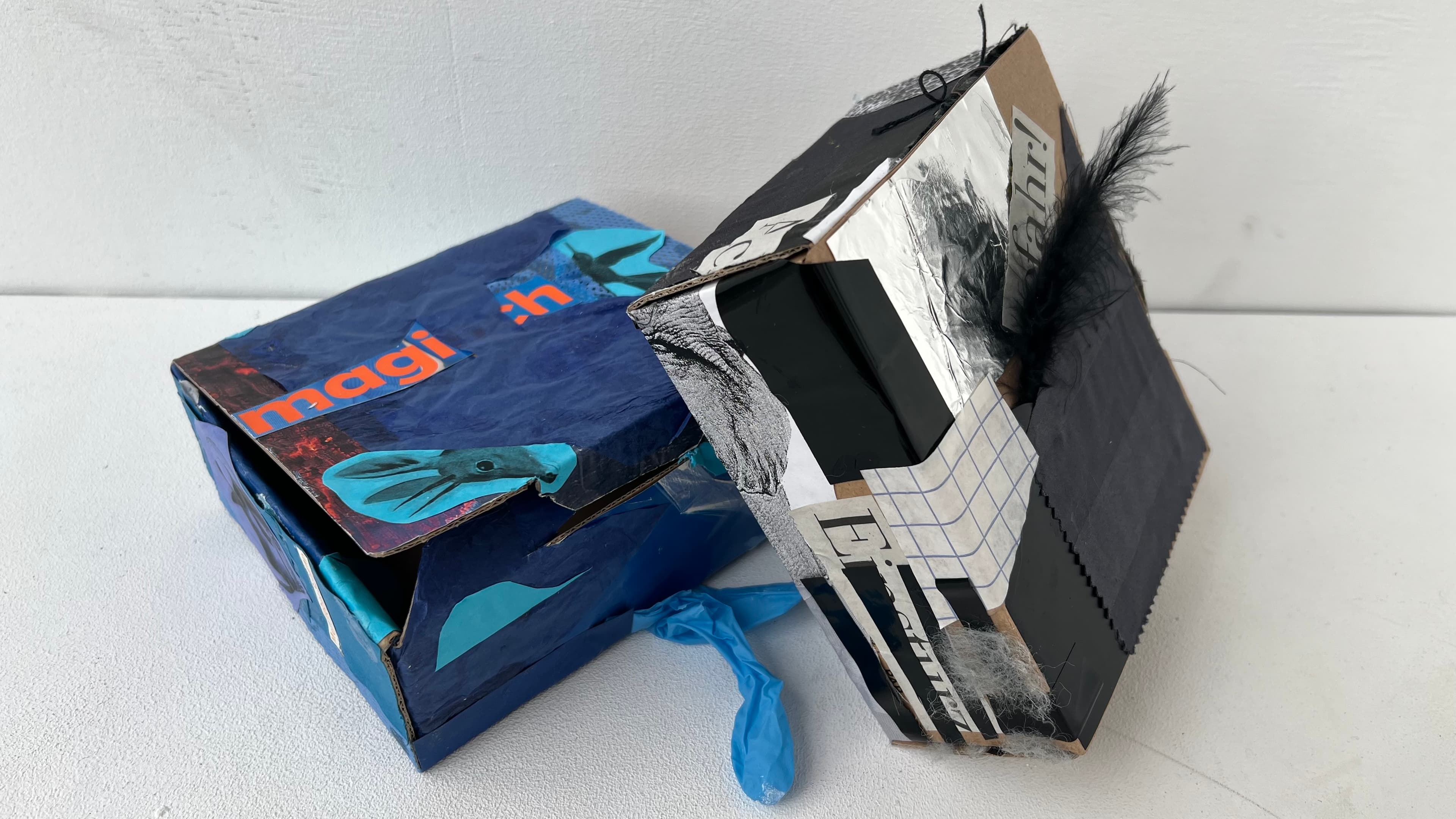 Zwei collagierte Kartonschachtel, eine in Blau und Orange, die andere in Schwarz und Silber mit einer Feder.